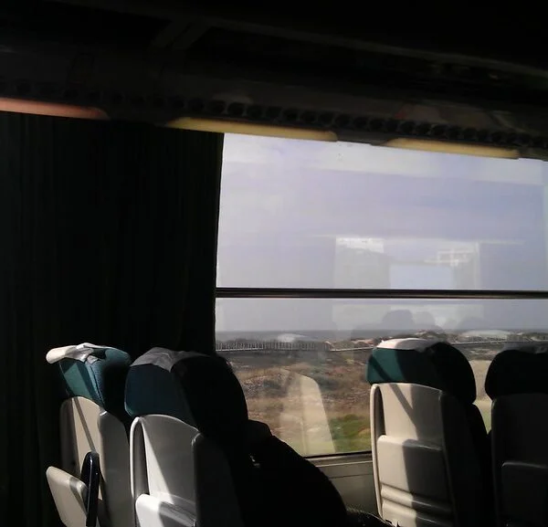 Interior of a bus with empty seats and green curtains, sunlight streaming through the windows with a view of arid landscape outside.