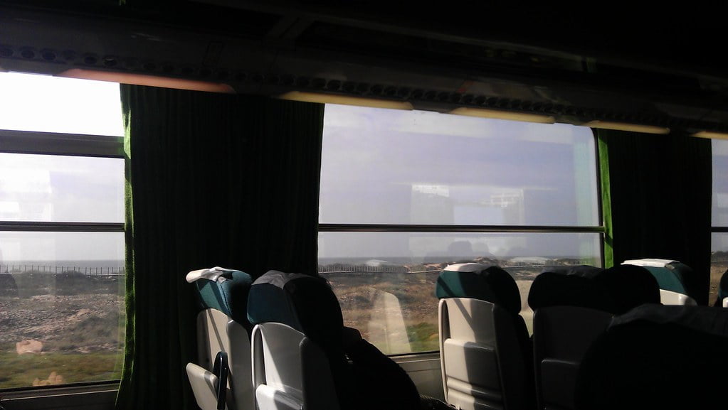 Interior of a bus with empty seats and green curtains, sunlight streaming through the windows with a view of arid landscape outside.