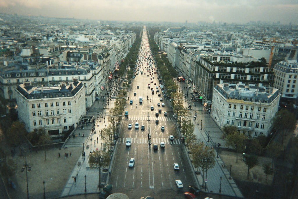 An aerial view of the Champs-Élysées in Paris, with traffic and classic Haussmann-style buildings lining the street, extending into the distance.