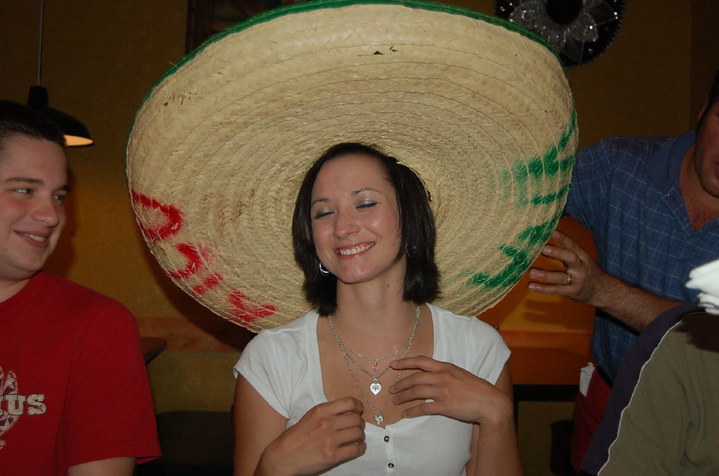A woman smiling with a large sombrero being placed on her head by someone out of frame, with two people visible beside her.