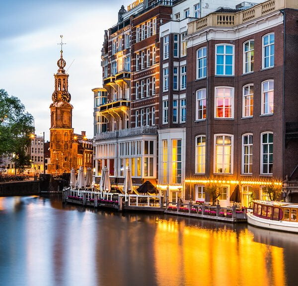 Twilight view of Amsterdam canals with illuminated buildings, a boat on the water, and the Munttoren (Mint Tower) in the background.