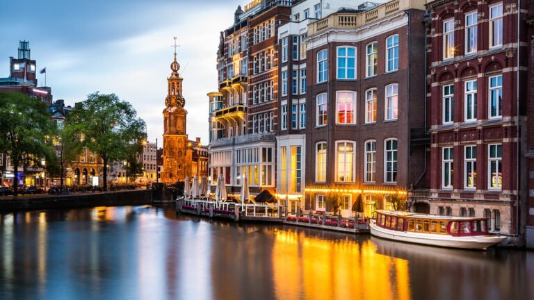 Twilight view of Amsterdam canals with illuminated buildings, a boat on the water, and the Munttoren (Mint Tower) in the background.