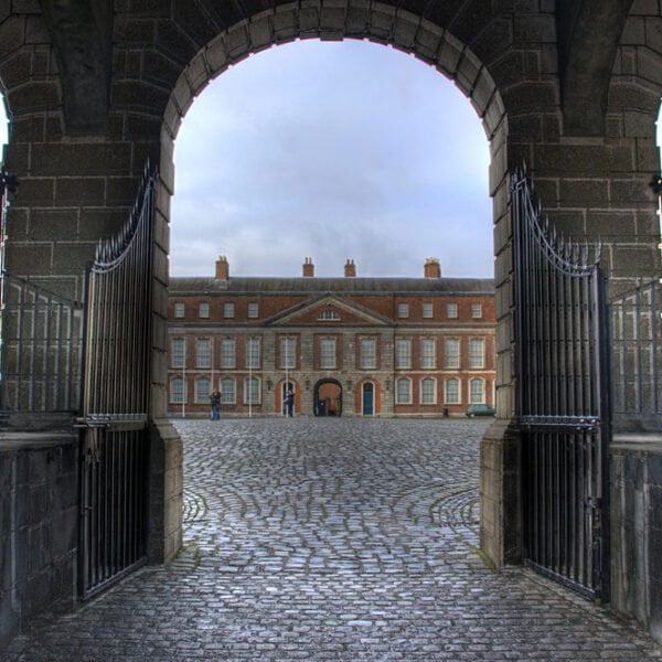 View through an archway with open wrought-iron gates leading to a cobbled courtyard and a classic red brick building facade with symmetrical windows.