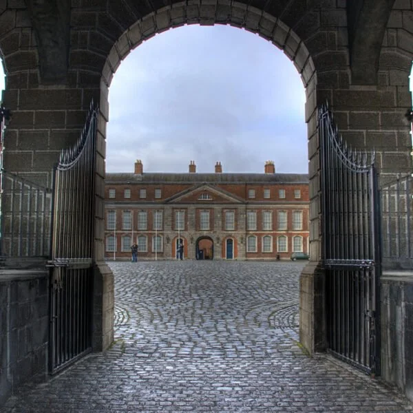 View through an archway with open wrought-iron gates leading to a cobbled courtyard and a classic red brick building facade with symmetrical windows.