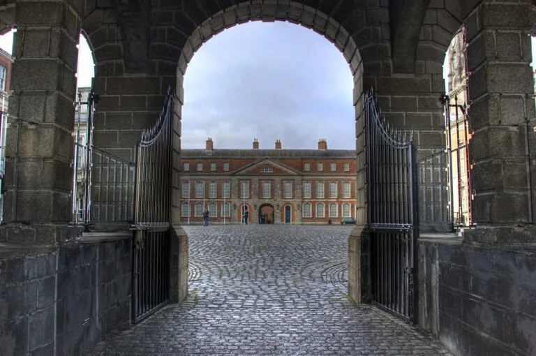 View through an archway with open wrought-iron gates leading to a cobbled courtyard and a classic red brick building facade with symmetrical windows.