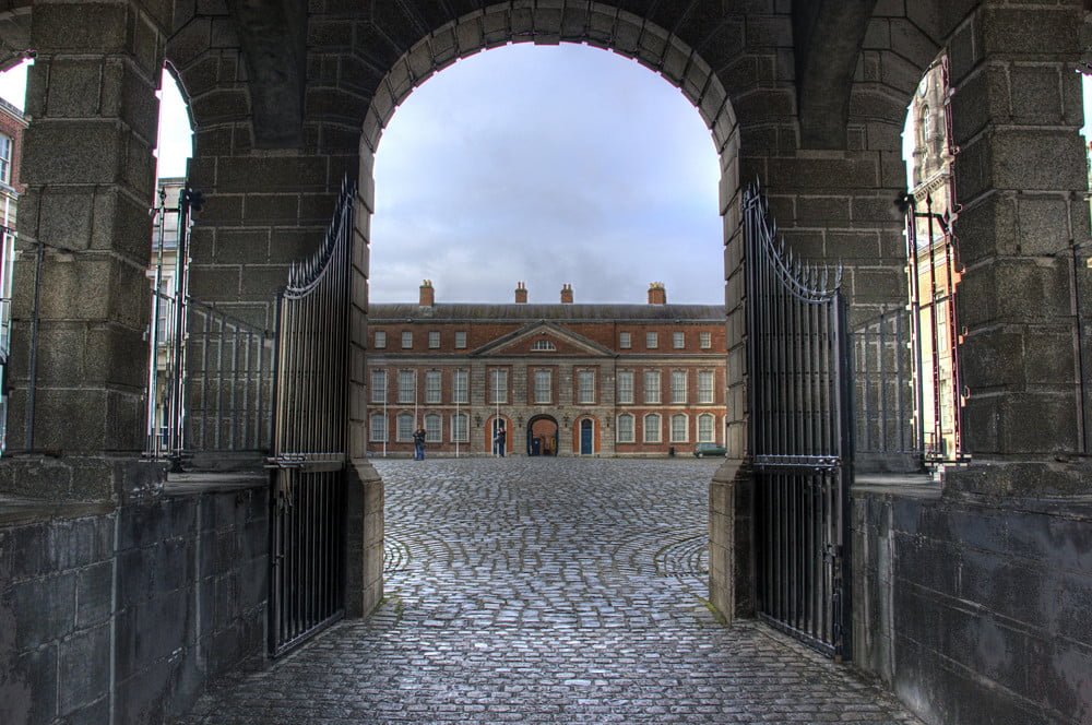 View through an archway with open wrought-iron gates leading to a cobbled courtyard and a classic red brick building facade with symmetrical windows.