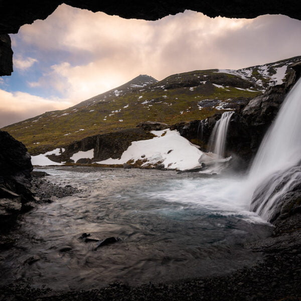 View from inside a cave of a waterfall cascading into a river with snow patches on a mountain landscape in the background.