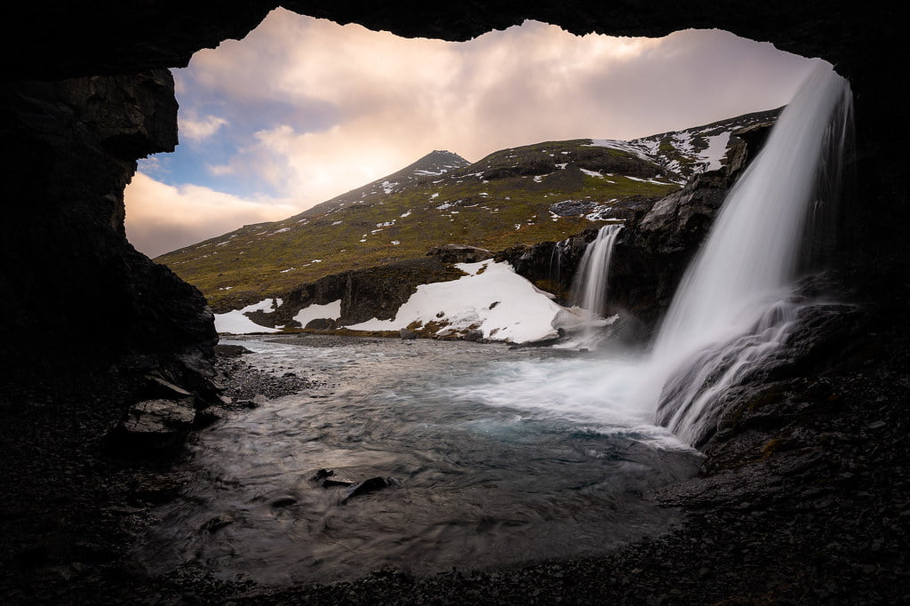 View from inside a cave of a waterfall cascading into a river with snow patches on a mountain landscape in the background.