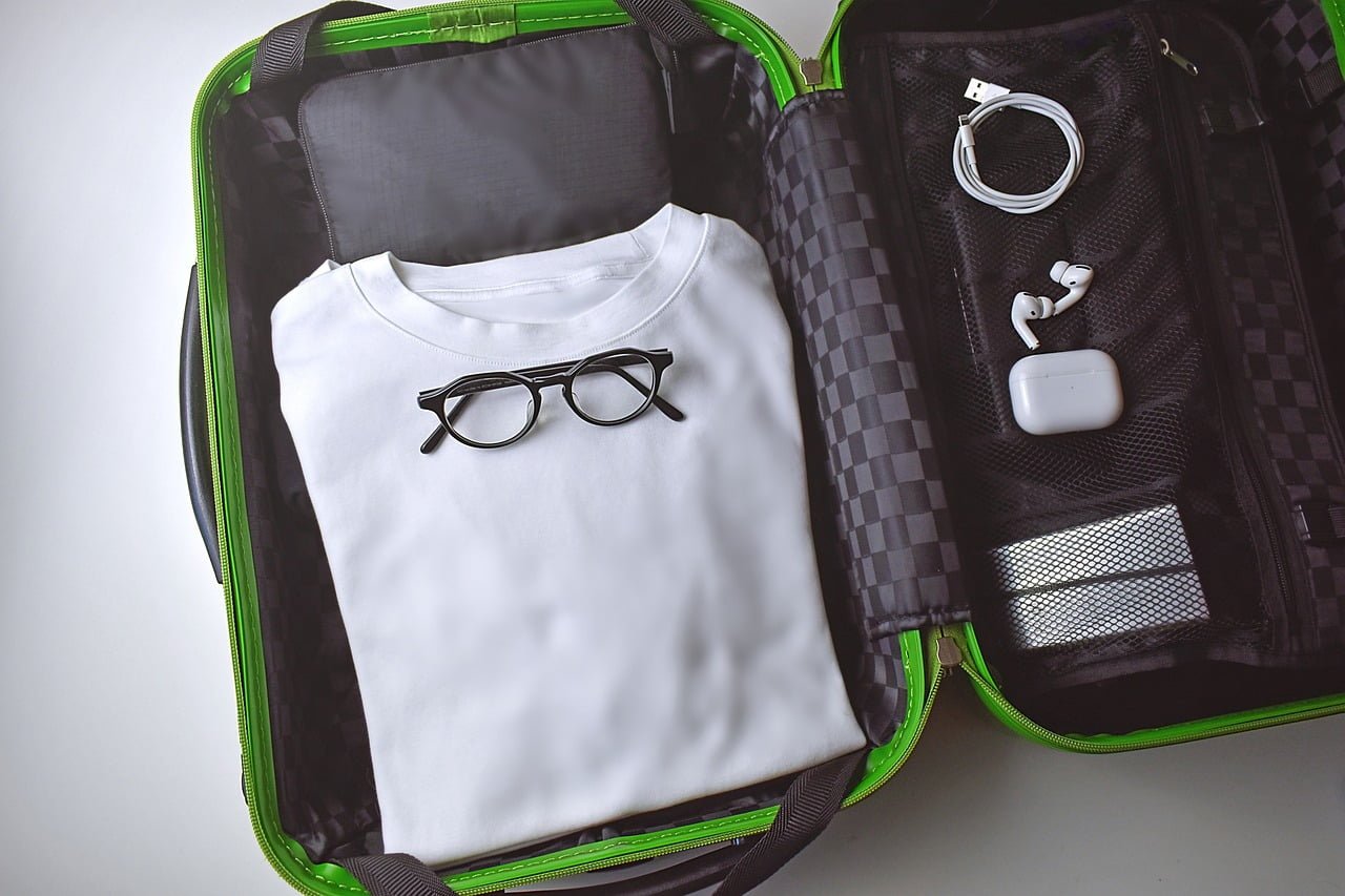 Open suitcase with neatly packed items including a folded white shirt, a pair of glasses, charging cable, and earphones in a black and green color scheme.