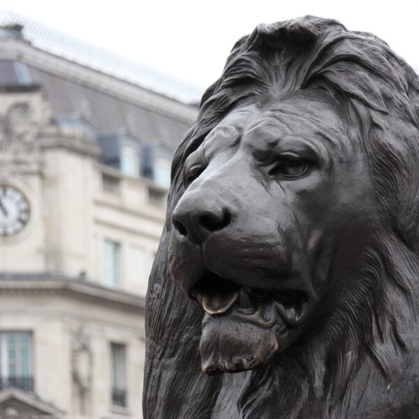 Close-up of a bronze lion sculpture with a blurred building and clock in the background.