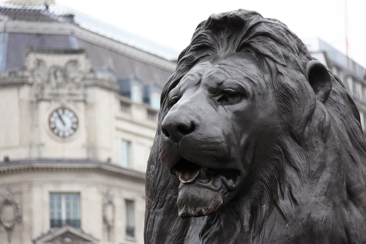 Close-up of a bronze lion sculpture with a blurred building and clock in the background.