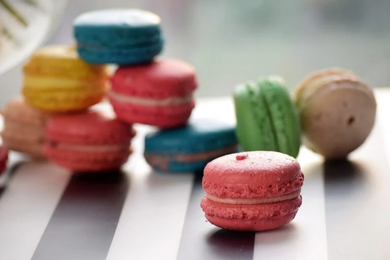 A selection of colorful macarons with a focus on a pink one in the foreground, displayed on a surface with alternating light and dark stripes.