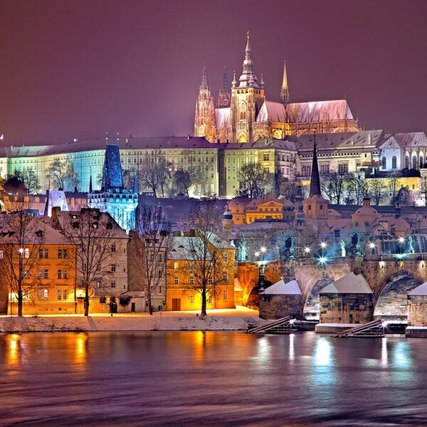 A night view of Prague showcasing the snow-covered Prague Castle and St. Vitus Cathedral, with the Charles Bridge in the foreground and the Vltava River partially illuminated by surrounding lights.