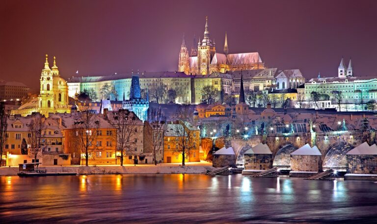 A night view of Prague showcasing the snow-covered Prague Castle and St. Vitus Cathedral, with the Charles Bridge in the foreground and the Vltava River partially illuminated by surrounding lights.