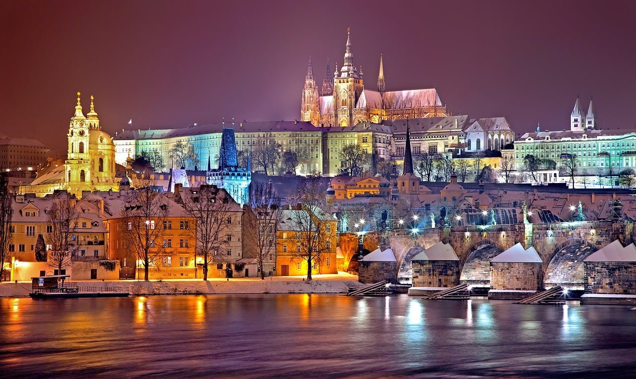 A night view of Prague showcasing the snow-covered Prague Castle and St. Vitus Cathedral, with the Charles Bridge in the foreground and the Vltava River partially illuminated by surrounding lights.