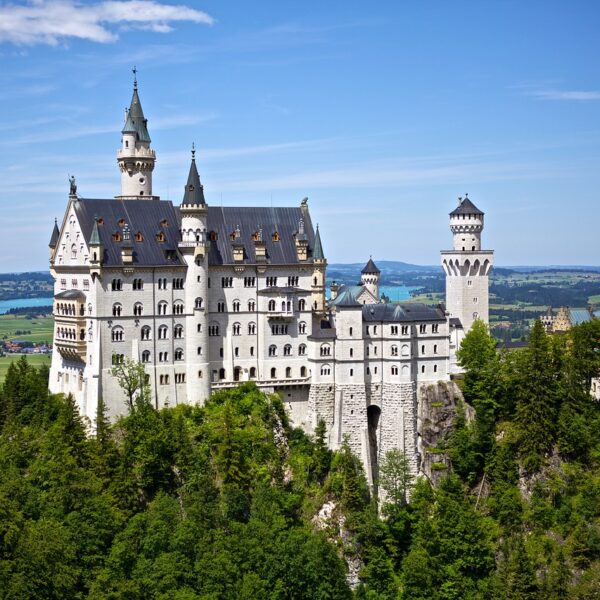 A picturesque view of the Neuschwanstein Castle perched on a lush hill with a backdrop of a lake and distant scenery under a clear blue sky.