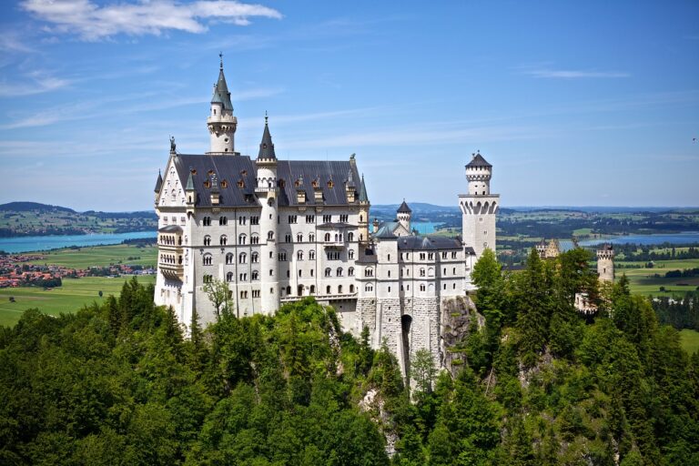 A picturesque view of the Neuschwanstein Castle perched on a lush hill with a backdrop of a lake and distant scenery under a clear blue sky.