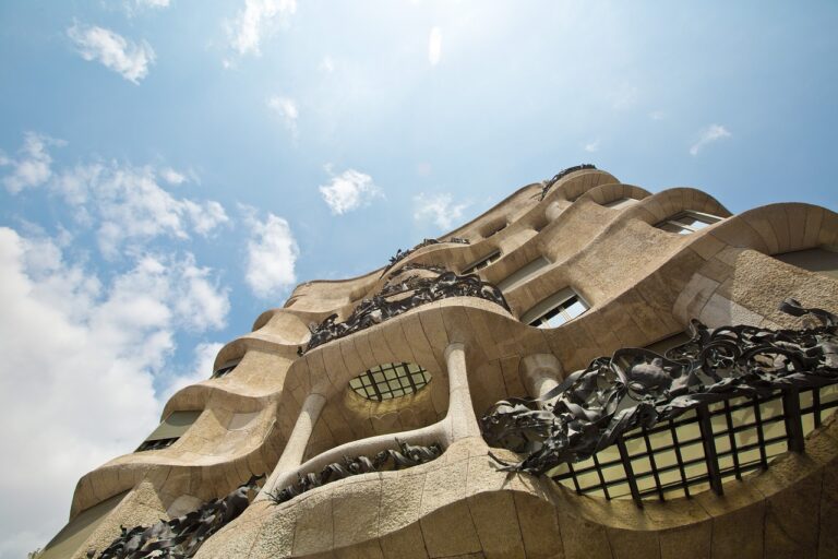 Exterior view of Casa Milà (La Pedrera) with its undulating stone facade and intricate wrought iron balconies against a blue sky with white clouds.