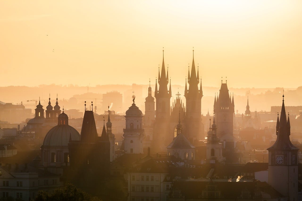 An aerial view of a city skyline at sunrise with silhouettes of historic buildings and spires against a golden sky.