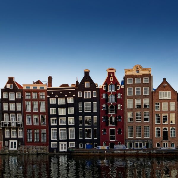 A row of traditional Dutch houses with gabled facades reflecting in the calm waters of a canal under a clear blue sky.