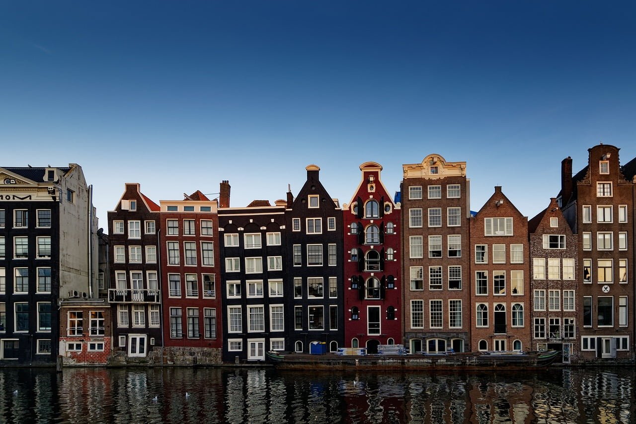 A row of traditional Dutch houses with gabled facades reflecting in the calm waters of a canal under a clear blue sky.
